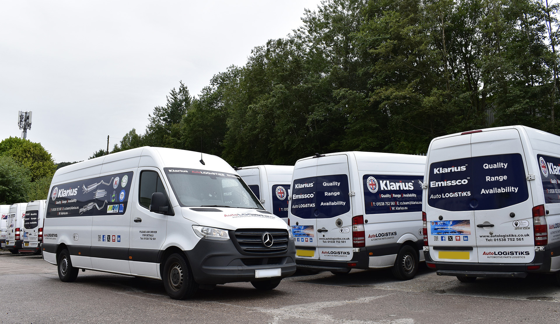 A fleet of Klarius branded white transit vans line up in a car park against a back drop of tall trees, with one van, closest to lense, driving behind a row of stationary vans facing the trees.